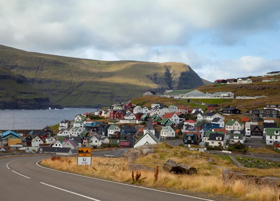 一條公路彎進法羅群島 Eiði 村,村裡彩色房舍沿著海灣分布,遠處是斷崖